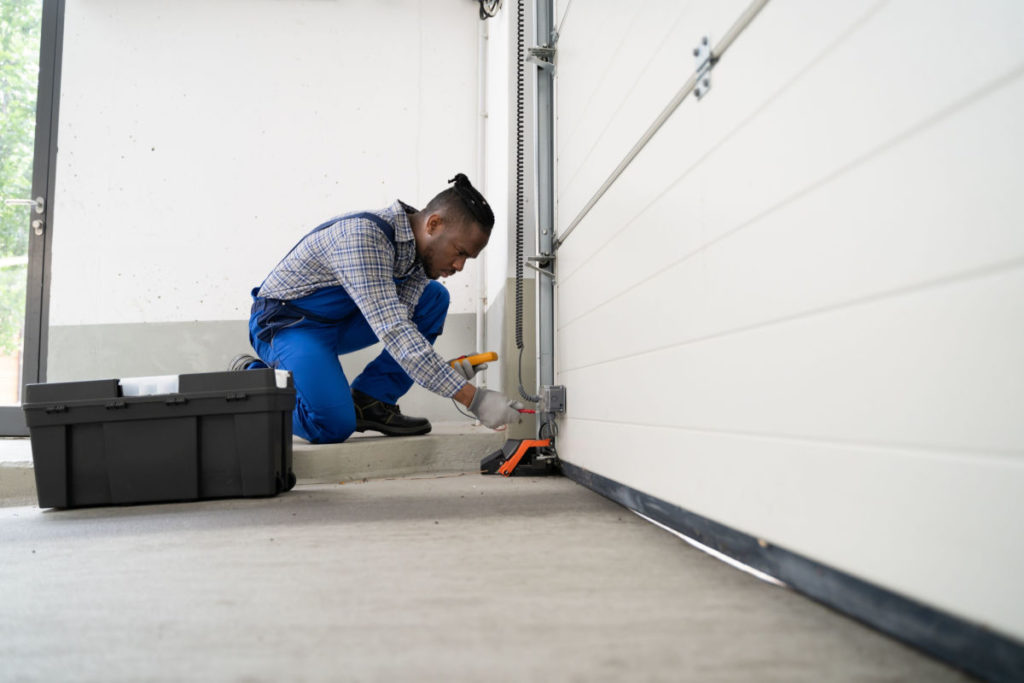 A man in blue overalls and a plaid shirt works on a garage door while kneeling.